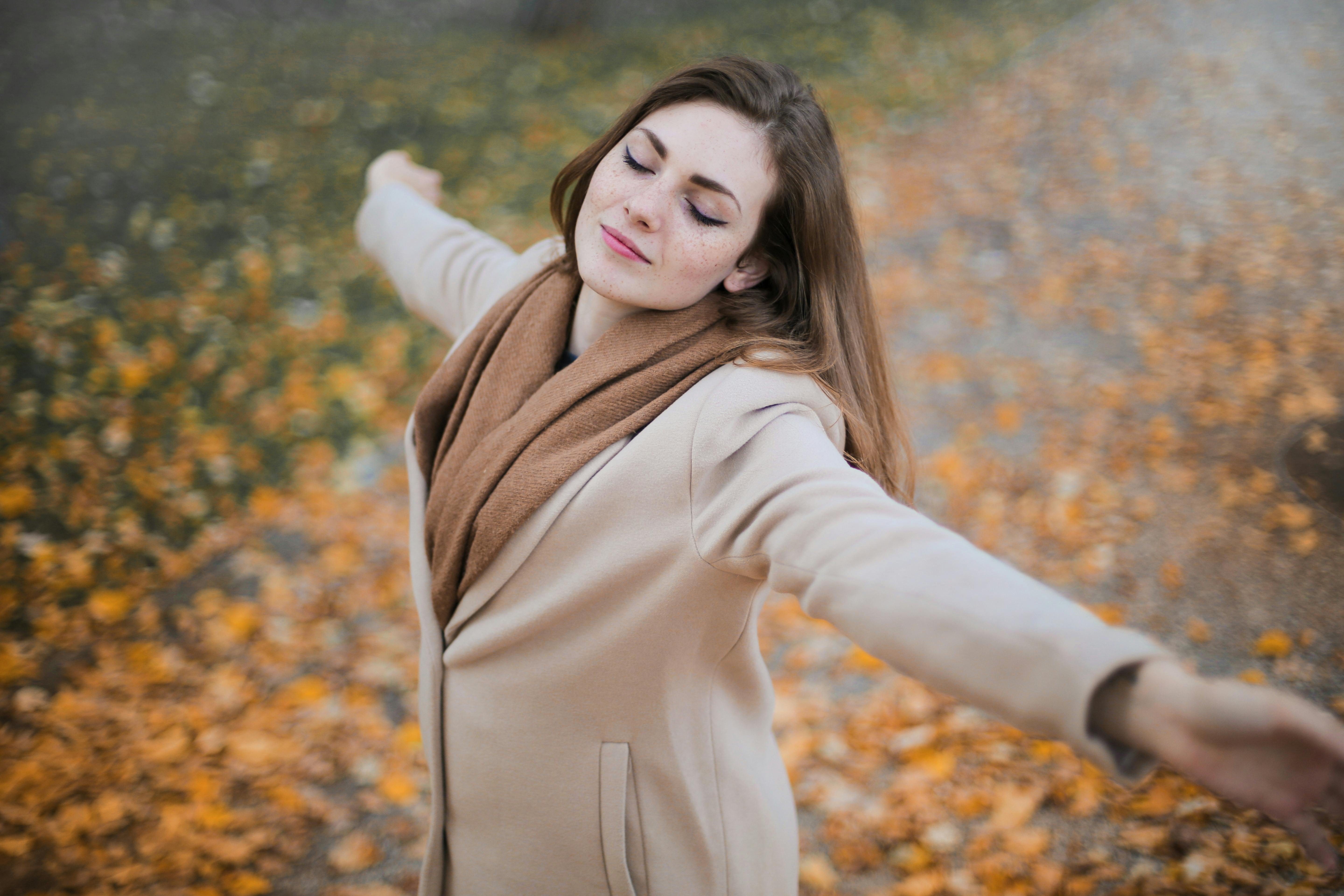 Woman in a beige coat and brown scarf standing in a field of autumn leaves.